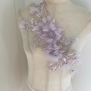 Puede incluir: Aplicación floral lavanda con intrincados abalorios, lentejuelas y delicados acentos de plumas. El diseño presenta múltiples racimos de flores y adornos en forma de hojas, creando un aspecto texturizado y elegante. La aplicación se muestra en un maniquí.