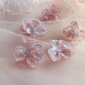 May include: Four individual light pink floral appliques crafted from delicate lace and embellished with shimmering sequins. Each flower features a layered petal design with a small white centre, creating a textured and reflective appearance.