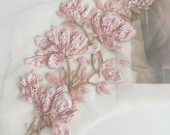 Può includere: Applicazione floreale con fiori e foglie di perline rosa. I fiori hanno più strati di petali e sono attaccati a un supporto in tessuto bianco trasparente. Le perline sono rosa chiaro e gli steli sono argentati.