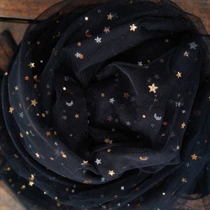 May include: Black tulle fabric with gold and silver star and crescent moon embellishments. The fabric is gathered and draped, creating a layered effect. The celestial design is scattered across the dark background.