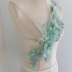 Peut inclure: Une applique vert d'eau clair avec des plumes, des paillettes et des perles. La décoration est disposée en forme de fleur. L'applique est présentée sur un mannequin. Cet article convient aux robes de mariée, de soirée ou autres vêtements de cérémonie.