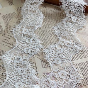 Puede incluir: Primer plano de un ribete de encaje blanco con un diseño floral. El encaje tiene un borde festoneado y se muestra sobre un fondo de documentos de papel vintage. El encaje es un textil decorativo delicado.