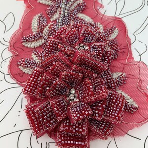 以下が含まれることがあります： 手作りの赤いビーズフラワーアップリケのクローズアップ。花は小さな正方形のビーズでできており、透明な赤い布の裏地に付いています。花の中心には透明なラインストーンのクラスターがあり、銀色の刺繍で囲まれています。