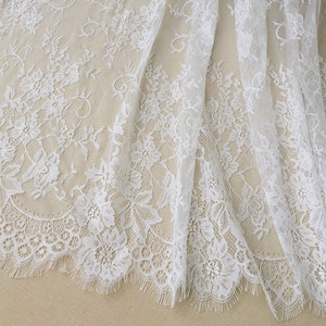 May include: White lace fabric with a delicate floral pattern. The lace has a scalloped edge and a soft, flowing texture.