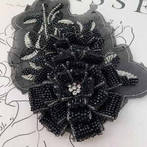 Könnte beinhalten: Schwarze, mit Perlen besetzte Blumenapplikation mit einem zentralen Cluster aus klaren Strasssteinen. Die Applikation befindet sich auf einem grauen Spitzenträger mit weißen Stickakzenten. Das Wort "MATISSE" ist im Hintergrund zu sehen.