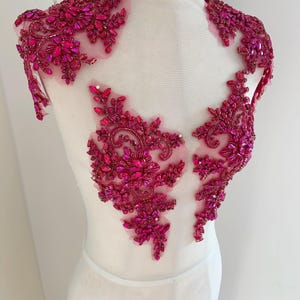 May include: A magenta beaded applique with intricate floral and geometric patterns. The embellishment features a high neckline and shoulder details, with a sheer mesh backing. The design is densely covered in sparkling beads and crystals, creating a luxurious look.