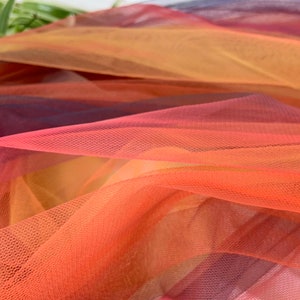 Op de afbeelding: Een close-up van een doorschijnende, ombre stof met een gradiënt van kleuren van donkerpaars naar oranje. De stof is zacht en delicaat, met een fijne gaastextuur.