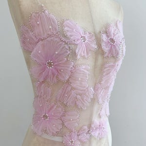May include: A sheer bodice applique featuring layered light pink fabric flowers with ruffled petals, accented by small white pearls and delicate silver beadwork, arranged in a floral pattern on a transparent mesh base on a dress form.