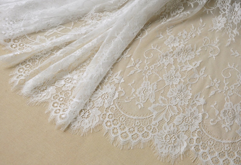 May include: White lace fabric with a delicate floral pattern. The lace has a scalloped edge and a soft, flowing texture.