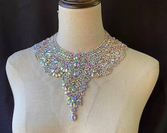 May include: A large, iridescent, crystal-encrusted statement necklace with a teardrop pendant. The necklace is made of many small, faceted crystals that are arranged in a cascading pattern.