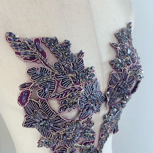 May include: Purple beaded floral applique with gold thread embroidery. This delicate embellishment features a cluster of flowers and leaves, perfect for adding a touch of elegance to clothing or crafts.