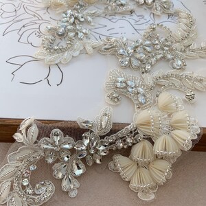 May include: An ivory lace appliqué featuring intricate floral patterns, embellished with clear faceted crystals, silver beads, and pleated fabric flower clusters. The design is arranged on a sheer mesh backing, showcasing detailed embroidery and sparkling accents.