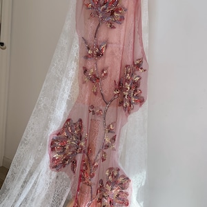 May include: A sheer, dusky pink fabric featuring embroidered floral designs in red, gold, and purple sequins. The fabric is layered over white lace, creating a delicate, textured effect. The floral embroidery is detailed and eye-catching, suitable for dressmaking.