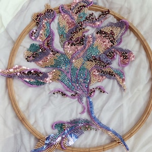 May include: A circular wooden embroidery hoop containing a floral design crafted with intricate beadwork and sequins in shades of lavender, teal, gold, and deep burgundy. The embroidery is worked onto a sheer, light purple fabric base.