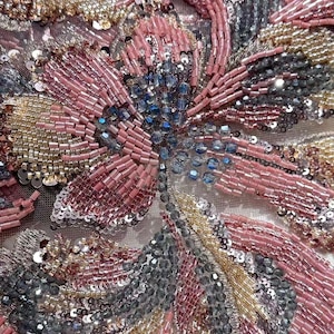 May include: Detailed close-up of a floral design, showcasing intricate beadwork and sequins. The design uses pink, silver, gold, and blue hues, creating a textured and shimmering effect. The beads are carefully arranged, forming a detailed pattern.
