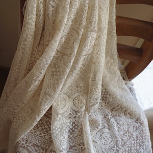 Puede incluir: Tela de encaje blanca con un delicado patr&oacute;n floral. La tela est&aacute; drapeada sobre una silla de madera.
