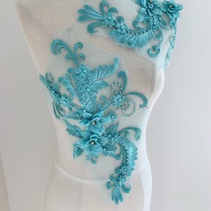 May include: A turquoise floral lace applique with intricate embroidered details. The applique features multiple flower designs, leaves, and swirling patterns. The lace is attached to a sheer, light blue fabric, creating a delicate and elegant look.