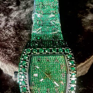 May include: A rectangular wristwatch with a green gemstone-encrusted band and face. The watch features a silver-colored crown and hands, with Roman numeral hour markers. The date is displayed at the 3 o'clock position.