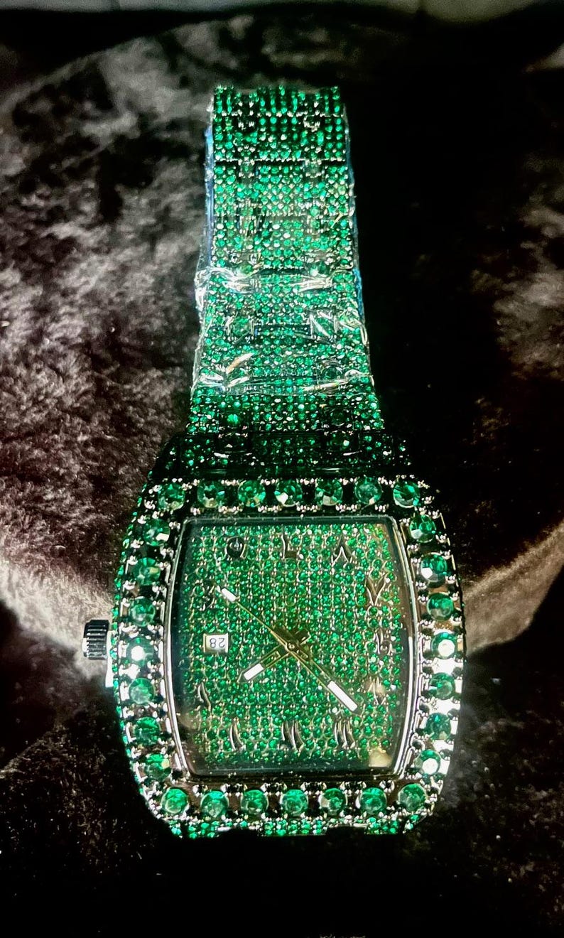 May include: A rectangular wristwatch with a green gemstone-encrusted band and face. The watch features a silver-colored crown and hands, with Roman numeral hour markers. The date is displayed at the 3 o'clock position.