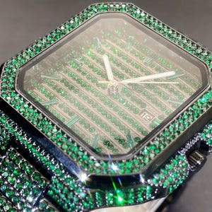 May include: A luxury wristwatch with a black case and band, lavishly encrusted with green gemstones. The watch face features a diamond-shaped design with green gems and silver hands. The date is displayed at the 3 o'clock position.