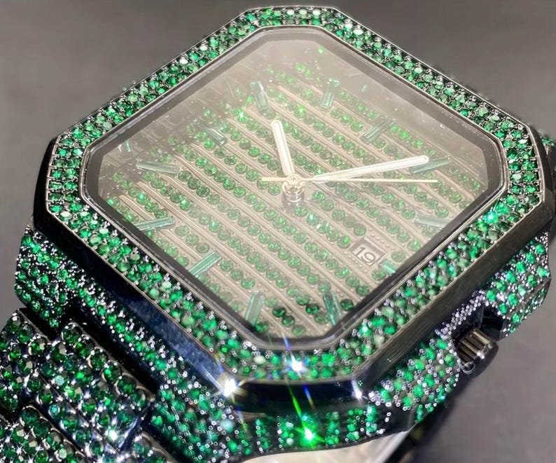 May include: A luxury wristwatch with a black case and band, lavishly encrusted with green gemstones. The watch face features a diamond-shaped design with green gems and silver hands. The date is displayed at the 3 o'clock position.