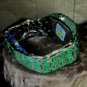 May include: A luxury wristwatch with a silver band and a face covered in small, sparkling green stones. The watch is displayed on a dark, textured surface, showcasing its intricate design and craftsmanship.