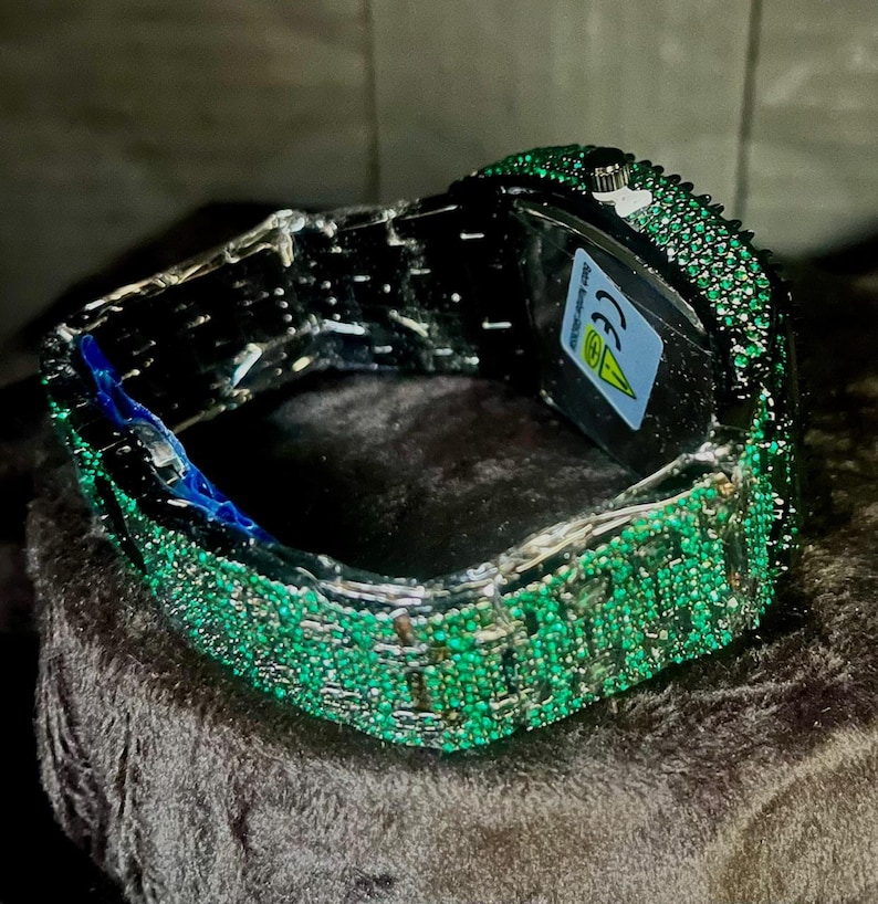May include: A luxury wristwatch with a silver band and a face covered in small, sparkling green stones. The watch is displayed on a dark, textured surface, showcasing its intricate design and craftsmanship.
