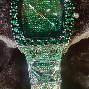 May include: A luxury wristwatch with a square green face and a silver band. The watch face is covered in small green stones, and the bezel and band are encrusted with larger green stones. The watch hands are silver, and the date is displayed as "28".