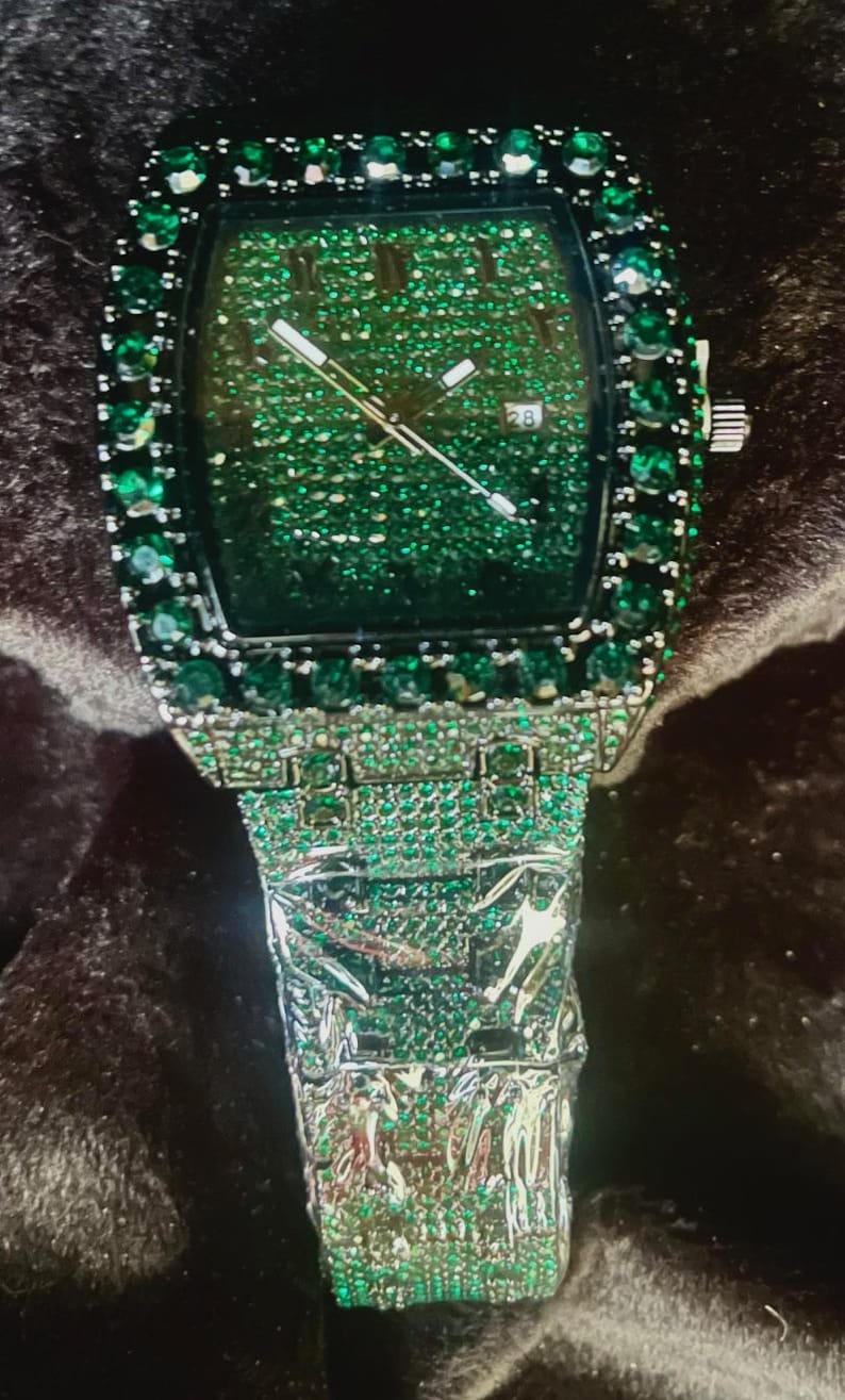 May include: A luxury wristwatch with a square green face and a silver band. The watch face is covered in small green stones, and the bezel and band are encrusted with larger green stones. The watch hands are silver, and the date is displayed as "28".