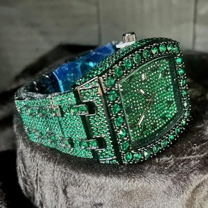 May include: A luxury wristwatch with a square green face and band, fully encrusted with small green gemstones. The watch has silver-colored metal accents and a classic watch clasp. The watch is displayed on a dark, textured surface.