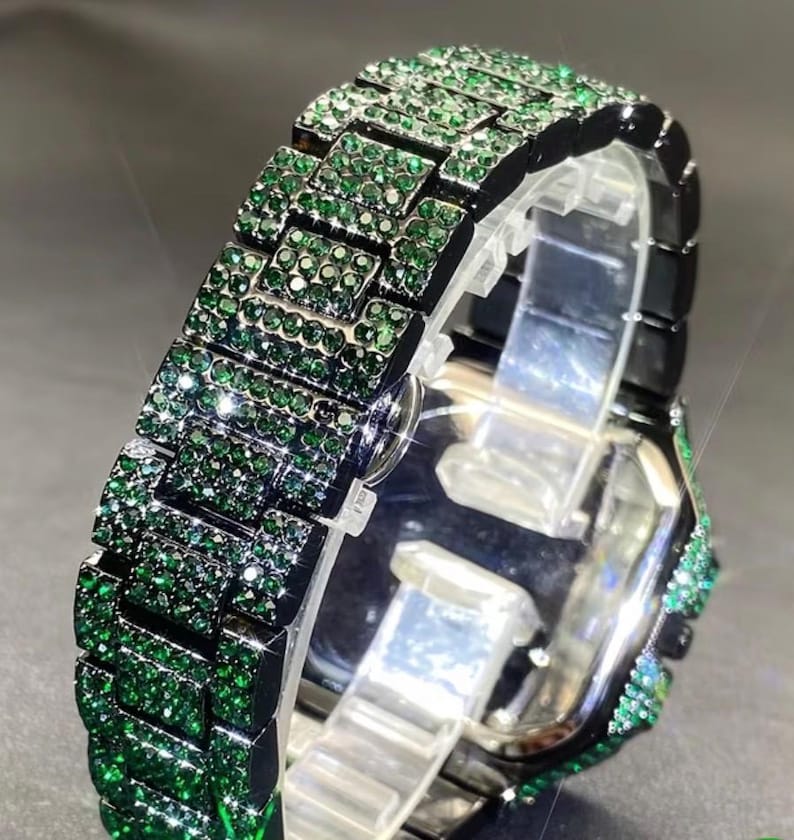 May include: A luxury watch band with a black frame and a dazzling array of emerald-green gemstones. The band is made of rectangular links, each covered in sparkling green stones. The watch face is silver, and the overall design is elegant and eye-catching.