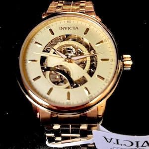 May include: A gold-toned Invicta watch with a skeleton dial, showcasing the intricate inner workings. The watch features a gold band and a round face with visible gears and hands. The watch is new and has a tag attached.