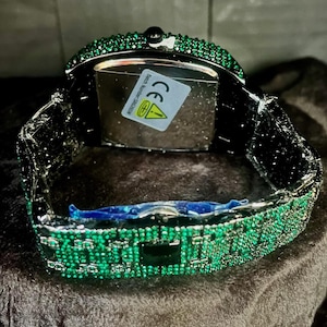 May include: A luxury wristwatch with a rectangular face and a band encrusted with green gemstones. The watch has a black metal frame and a clasp. The watch face is silver with a sticker on the back.