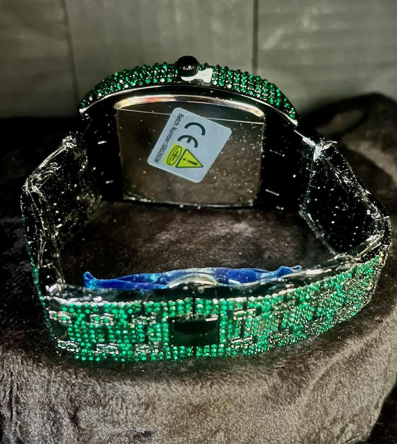 May include: A luxury wristwatch with a rectangular face and a band encrusted with green gemstones. The watch has a black metal frame and a clasp. The watch face is silver with a sticker on the back.