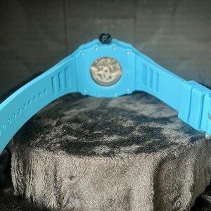 May include: A light blue wristwatch with a transparent watch face, showcasing the inner workings. The watch has a matching light blue band with a silver buckle. The watch is displayed on a dark, textured surface.