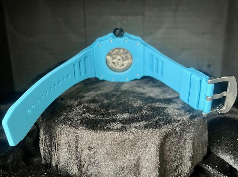 May include: A light blue wristwatch with a transparent watch face, showcasing the inner workings. The watch has a matching light blue band with a silver buckle. The watch is displayed on a dark, textured surface.