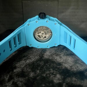 May include: A light blue wristwatch with a transparent back, showcasing the intricate inner workings. The watch has a matching light blue band with rectangular cutouts. The crown is black.