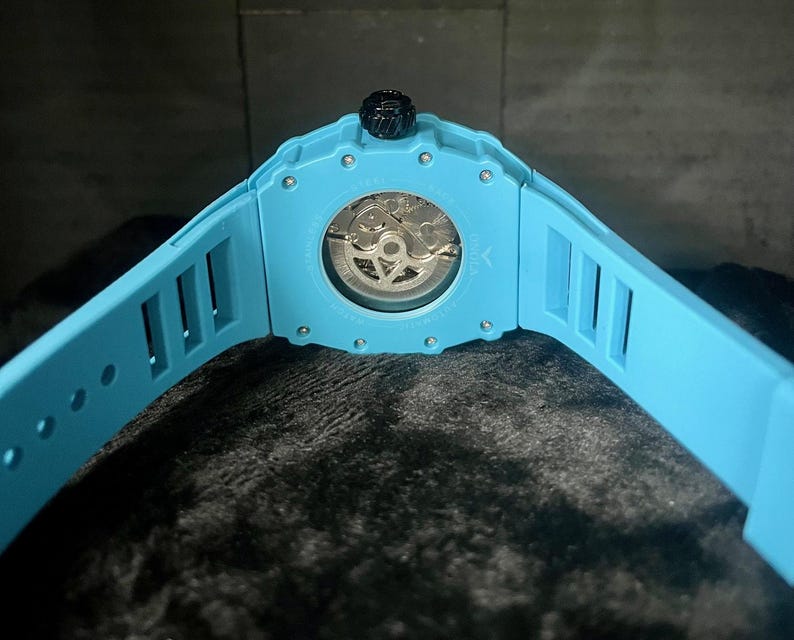 May include: A light blue wristwatch with a transparent back, showcasing the intricate inner workings. The watch has a matching light blue band with rectangular cutouts. The crown is black.
