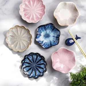 Puede incluir: Un conjunto de platos de cerámica con forma de flor en varios colores, incluyendo rosa, azul y blanco. Los platos tienen superficies texturizadas y están dispuestos sobre una superficie de mármol. También hay un reposa palillos azul y palillos.