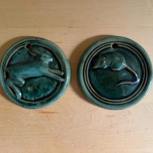 May include: Two circular ceramic wall plaques with a glossy teal glaze. One plaque features a relief sculpture of a leaping rabbit, while the other displays a relief sculpture of a mouse. Each piece has a small hole at the top for hanging.