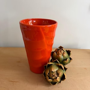 May include: A bright orange ceramic tumbler with a textured, rippled surface. Next to the tumbler are two decorative artichoke ornaments, each featuring layered, pointed leaves in shades of muted green and brown with subtle yellow highlights.