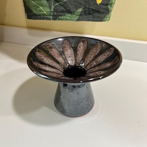 May include: A ceramic pedestal bowl with a dark gray base and a wide, shallow top. The top features a dark brown and tan floral pattern radiating from a central black opening. The bowl is likely handmade and suitable for decorative purposes.