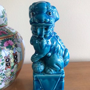 May include: A turquoise ceramic Foo Dog statue. The dog is seated on a square base, holding a ball. The statue has detailed features, including a textured mane and open mouth. The statue is a decorative item.