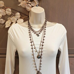 May include: Three-strand silver-toned necklace set with turquoise-colored stones and small beads. The necklaces are displayed on a white mannequin, with a white long-sleeved shirt. The background includes a wooden wall and decorative leaves.