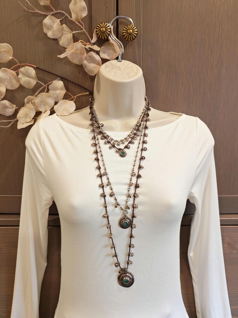 May include: Three-strand silver-toned necklace set with turquoise-colored stones and small beads. The necklaces are displayed on a white mannequin, with a white long-sleeved shirt. The background includes a wooden wall and decorative leaves.