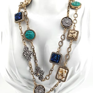 May include: A gold-toned necklace with multiple strands. The necklace features various decorative elements, including turquoise-colored stones, square blue glitter stones, and floral and cross-shaped accents. The necklace is displayed on a white mannequin.