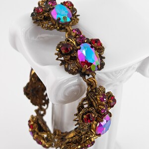May include: Vintage-style bracelet with ornate gold-tone metalwork. The bracelet features large, oval-shaped iridescent stones in shades of blue, purple, and pink, surrounded by smaller red and pink gemstones. A classic piece of jewelry.