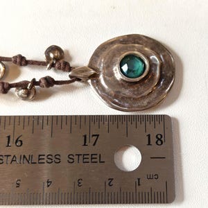 May include: A silver pendant necklace with a large, round, textured pendant featuring a central, emerald-green gemstone. The pendant is connected to a brown cord with small silver bells. The ruler shows the pendant is approximately 1.5 inches in diameter.