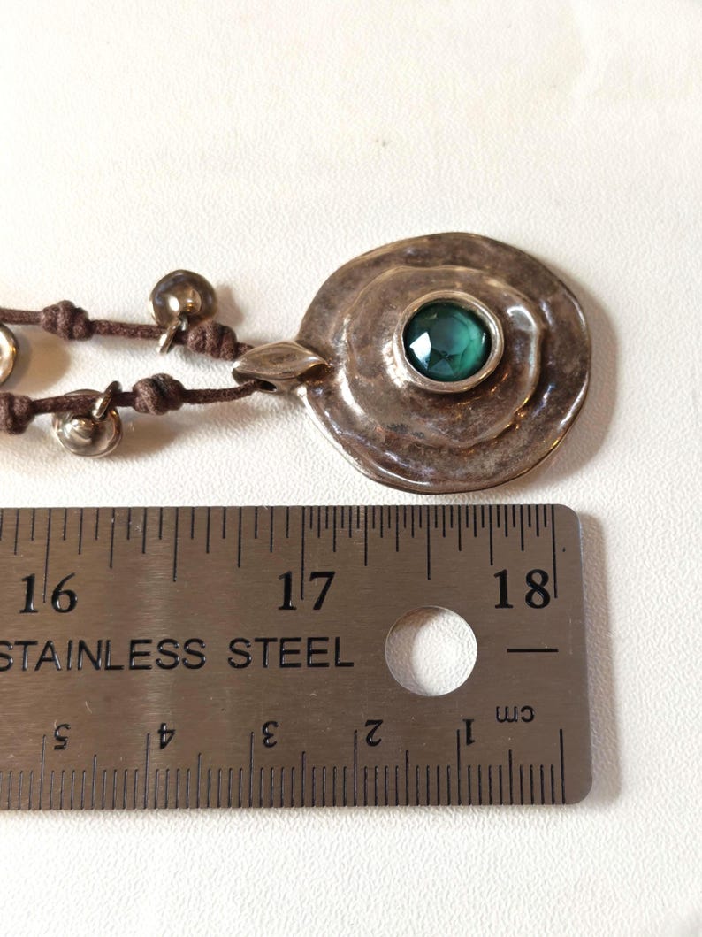 May include: A silver pendant necklace with a large, round, textured pendant featuring a central, emerald-green gemstone. The pendant is connected to a brown cord with small silver bells. The ruler shows the pendant is approximately 1.5 inches in diameter.
