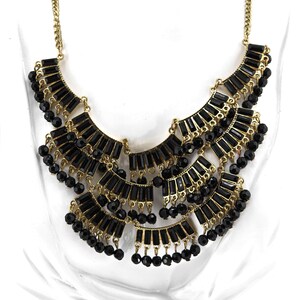 May include: A statement necklace featuring a tiered design with black rectangular and round beads. The necklace has a gold-tone chain and accents, creating a bold and elegant look. The necklace is designed to be worn around the neck.